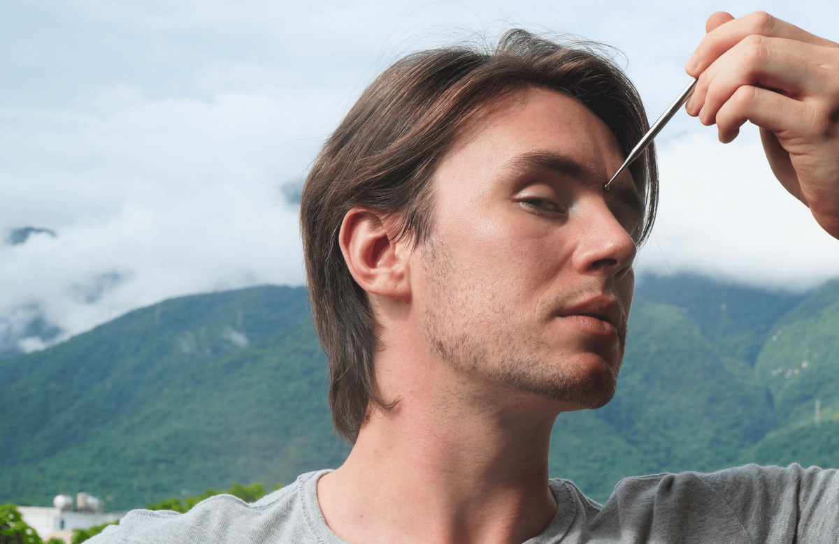 A close-up lifestyle shot of a man outdoors with mountains and clouds in the background, holding a small metal tool near his eyebrow.